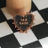 This Sad Babe Copper pin features a barbed wire heart and traditional tattoo design, perfect for adding an emo touch to your pin collection.