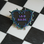 This Sad Babe pin features a barbed wire heart in rainbow metal, blending traditional tattoo style. 