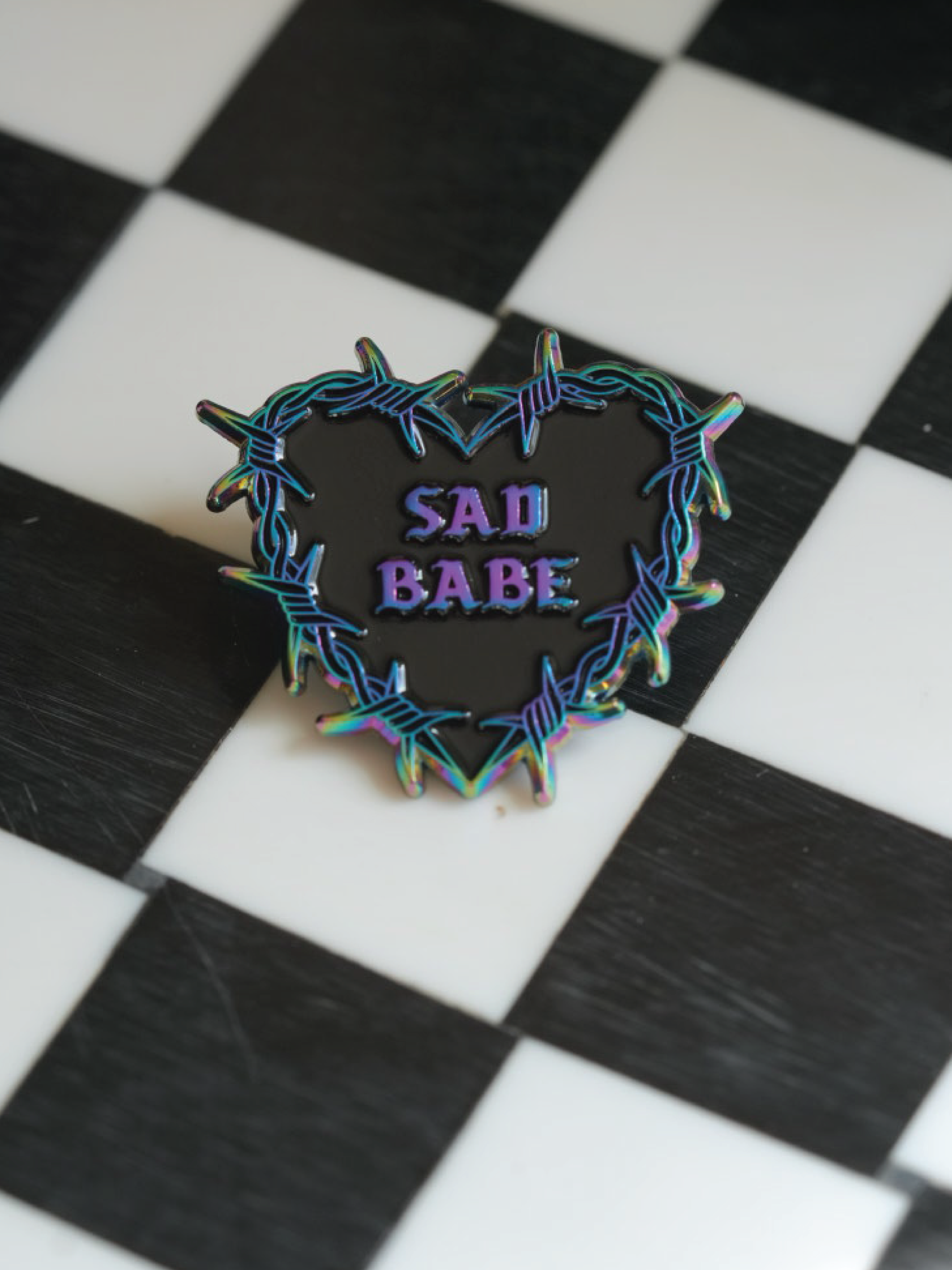This Sad Babe pin features a barbed wire heart in rainbow metal, blending traditional tattoo style. 