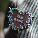 Perfect addition to any emo pin collection, this Sad Babe pin showcases a rainbow metal barbed wire heart and traditional tattoo style.