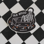 Close-up of the 'Sad Diego Gothic' sticker with a white skeleton lying in a coffin. Sticker is on a black and white checkered background.