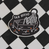 Close-up of the 'Sad Diego Gothic' sticker with a white skeleton lying in a coffin. Sticker is on a black and white checkered background.