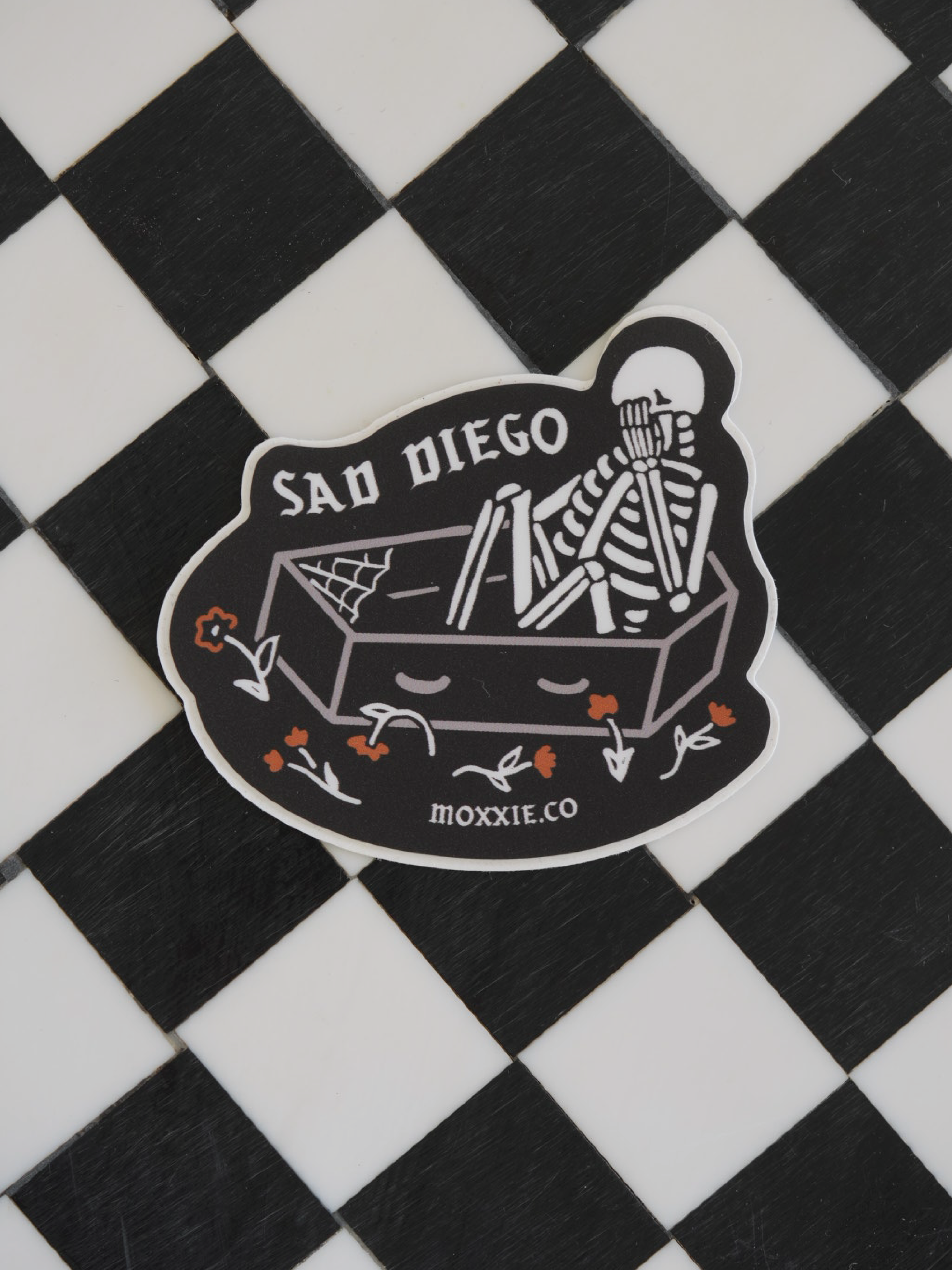 Close-up of the 'Sad Diego Gothic' sticker with a white skeleton lying in a coffin. Sticker is on a black and white checkered background.